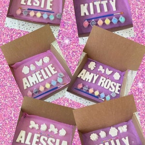 May include: Five open boxes with purple tissue paper inside. Each box contains a set of white ceramic letters spelling out a name, along with small white ceramic decorations and a set of paint pots. The names are Elsie, Kitty, Amelie, Amy Rose, and Alessia.