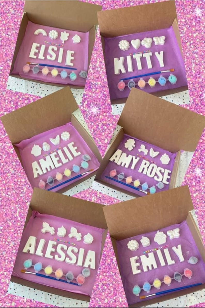 May include: Five open boxes with purple tissue paper inside. Each box contains a set of white ceramic letters spelling out a name, along with small white ceramic decorations and a set of paint pots. The names are Elsie, Kitty, Amelie, Amy Rose, and Alessia.