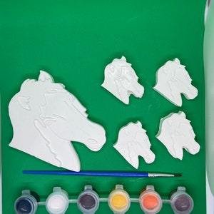 May include: A set of five white ceramic horse head figurines with a paintbrush and six small paint pots. The paint pots are black, white, grey, yellow, orange, and brown.