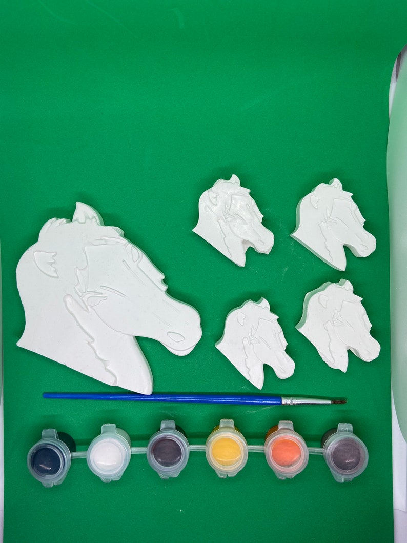 May include: A set of five white ceramic horse head figurines with a paintbrush and six small paint pots. The paint pots are black, white, grey, yellow, orange, and brown.
