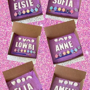 May include: Five boxes with purple tissue paper inside. Each box contains a set of white ceramic letters spelling out a name, paint pots, and a paintbrush. The names are Elsie, Sofia, Lowri, Anne, and El La.