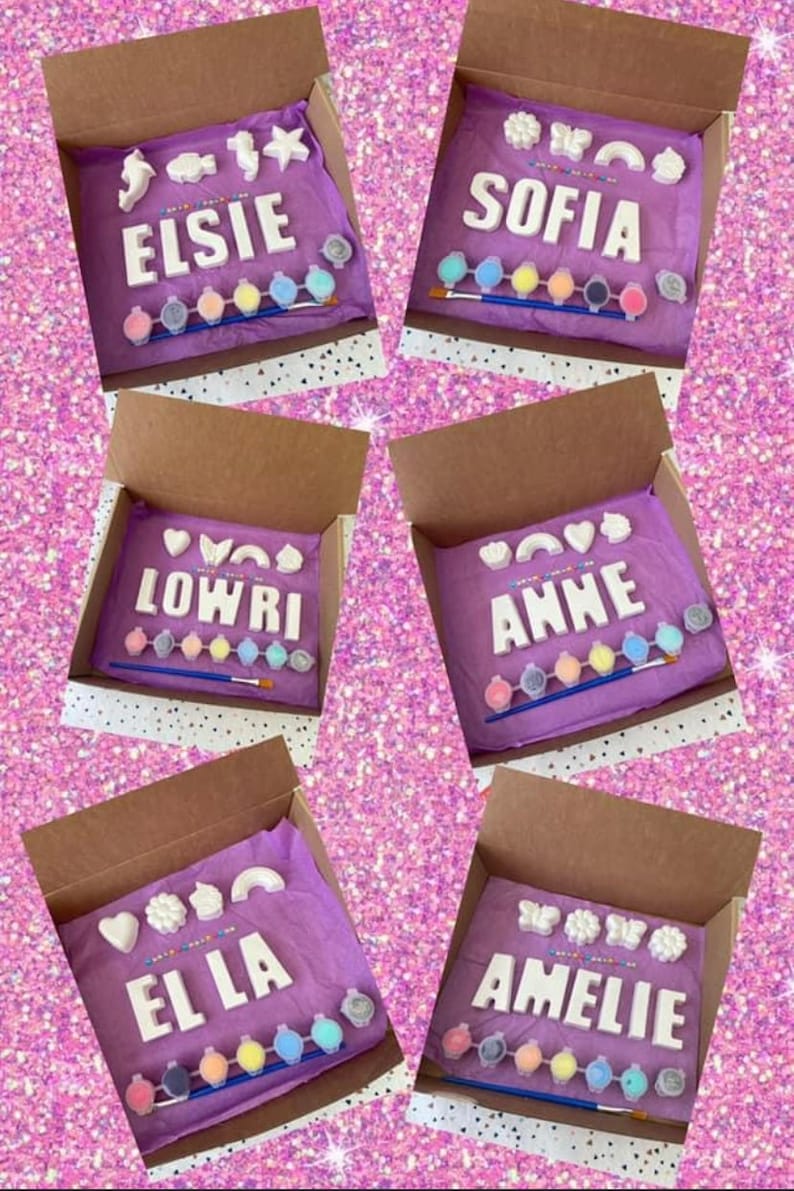 May include: Five boxes with purple tissue paper inside. Each box contains a set of white ceramic letters spelling out a name, paint pots, and a paintbrush. The names are Elsie, Sofia, Lowri, Anne, and El La.