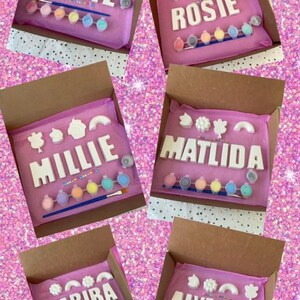 May include: Five pink boxes with white letters spelling out names, each box contains paint pots and a paintbrush. The names are Billie, Rosie, Millie, Matilda, and Habiba.