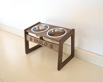May include: A dark brown wooden elevated pet feeder stand featuring two circular stainless steel bowls filled with dry food. The front panel is engraved with the name PABLO flanked by two paw print icons on either side of the text.