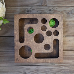 May include: A wooden pet puzzle toy with a variety of cutouts and holes. The toy is brown and has a small green ball inside one of the holes.