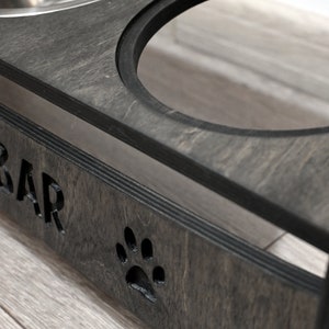 May include: A dark grey wooden pet food bowl stand with the words "Food Bar" engraved on the front. The stand has two stainless steel bowls and a paw print cutout.