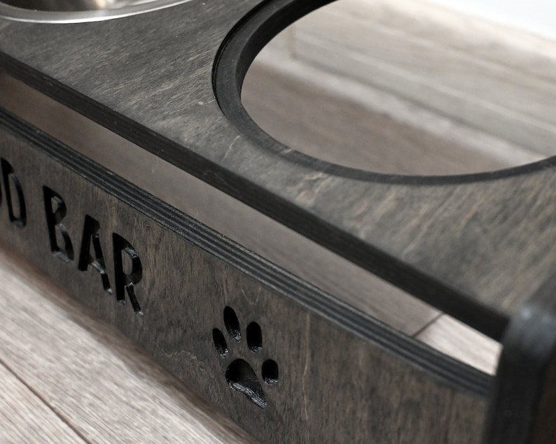 May include: A dark grey wooden pet food bowl stand with the words "Food Bar" engraved on the front. The stand has two stainless steel bowls and a paw print cutout.