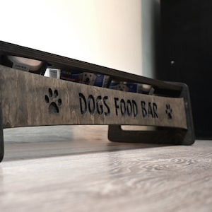 May include: A wooden dog food station with the words "Dogs Food Bar" carved into the front. The station has two paw prints carved on either side of the text. The station is stained a dark brown colour.