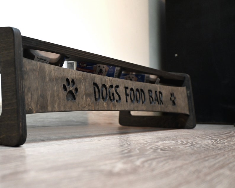 May include: A wooden dog food station with the words "Dogs Food Bar" carved into the front. The station has two paw prints carved on either side of the text. The station is stained a dark brown colour.