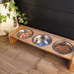 May include: A wooden pet food stand with three stainless steel bowls. The stand is made of light brown wood and has a natural finish. The bowls are filled with different types of pet food.