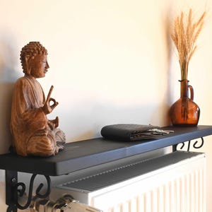 May include: A wooden Buddha statue sits on a black shelf, next to a dark wallet and a brown glass vase with wheat stalks. The shelf is mounted above a white radiator, with decorative black metal brackets.