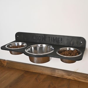 Könnte beinhalten: Ein schwarzer, an der Wand befestigter Futternapf mit drei Edelstahlnäpfen. Auf dem Holzregal steht der Schriftzug "CHOW TIME!". Jede Schüssel ist mit Trockenfutter gefüllt.