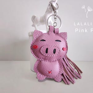 May include: A pink pig keychain made of leather with a fringe tassel. The pig has a stitched snout and eyes, and red felt cheeks. The keychain is attached to a silver key ring.