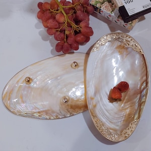 May include: Two oval-shaped serving dishes made of mother of pearl with gold trim. The dishes have a pearlescent white and gold color. The larger dish has a decorative design around the rim. The smaller dish has three gold feet. The dishes are on a white surface.