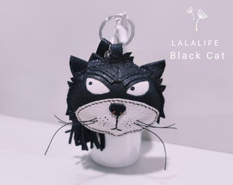 May include: Black leather cat keychain with white felt details. The cat has a grumpy expression and a long, black tassel.