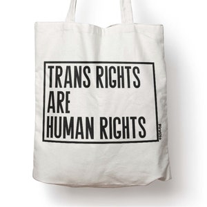 May include: White canvas tote bag with black text that reads "Trans Rights Are Human Rights" in a black box. The text "Piratito" is printed in black on the bottom right corner of the bag.