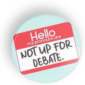 May include: A round, blue button with a red name tag that says "Hello my pronouns are NOT UP FOR DEBATE."