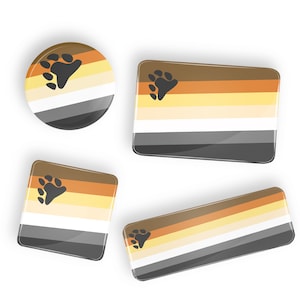 Peut inclure: Quatre épingles de drapeau de la fierté de l'ours de différentes tailles. Les épingles sont brunes, beiges, blanches et grises avec une empreinte de patte noire dans le coin supérieur gauche.