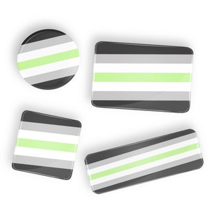 May include: Four different sizes of an asexuality pride flag buttons. The flag is black, grey, and light green with a light green stripe in the centre.