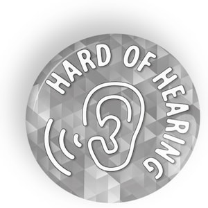 Puede incluir: Un botón redondo gris con las palabras "HARD OF HEARING" en blanco arqueadas en el borde. Un contorno blanco de una oreja con ondas sonoras está en el centro. El botón tiene un patrón geométrico.