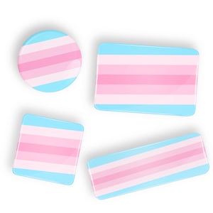 May include: Four different sized buttons with the transgender pride flag colors: light blue, pink, and light blue. The buttons are arranged in a square shape.