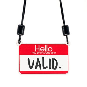 May include: A red name tag with a white border and black text that reads "Hello my pronouns are VALID." The name tag is hanging from a black lanyard with black beads.