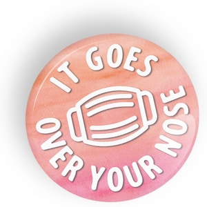 Puede incluir: Botón redondo con un fondo de estilo acuarela en tonos rosa y melocotón. El texto blanco dice "IT GOES OVER YOUR NOSE" alrededor de un contorno blanco de una mascarilla.