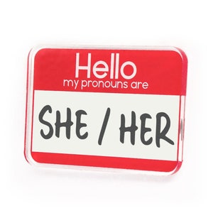 May include: Red name tag with white text that reads "Hello my pronouns are SHE / HER".
