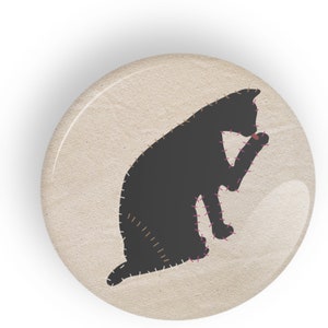 Op de afbeelding: Een ronde knoop met een zwart katten silhouet, gestikt met roze en bruin garen, op een beige achtergrond. De kat verzorgt zichzelf. De knoop heeft een glanzende, heldere afwerking.