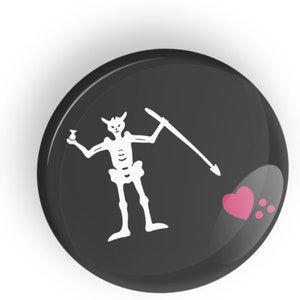Puede incluir: Un botón negro y blanco con un diseño de calavera de pirata y huesos cruzados. La calavera sostiene una botella y una lanza. Un corazón rosa se encuentra en la esquina inferior derecha.