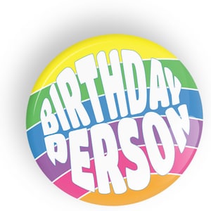 Op de afbeelding: Een ronde knop met een regenboogstreepontwerp. De tekst "BIRTHDAY PERSON" staat in het wit op de knop.