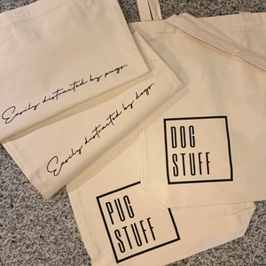 Op de afbeelding: Drie witte canvas tassen met zwarte tekst. De bovenste tas zegt "Gemakkelijk afgeleid door mopshonden." De middelste tas zegt "Gemakkelijk afgeleid door honden." De onderste tas zegt "PUG STUFF" in een zwart vierkant en "DOG STUFF" in een zwart vierkant.