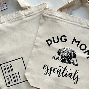 Op de afbeelding: Twee beige canvas tassen met zwarte tekst en afbeeldingen. E&eacute;n tas zegt "Pug Mom essentials" met een zwart-witte illustratie van een mops die een bloemenkrans draagt. De andere tas zegt "Pug Stuff" in een zwart vierkant.