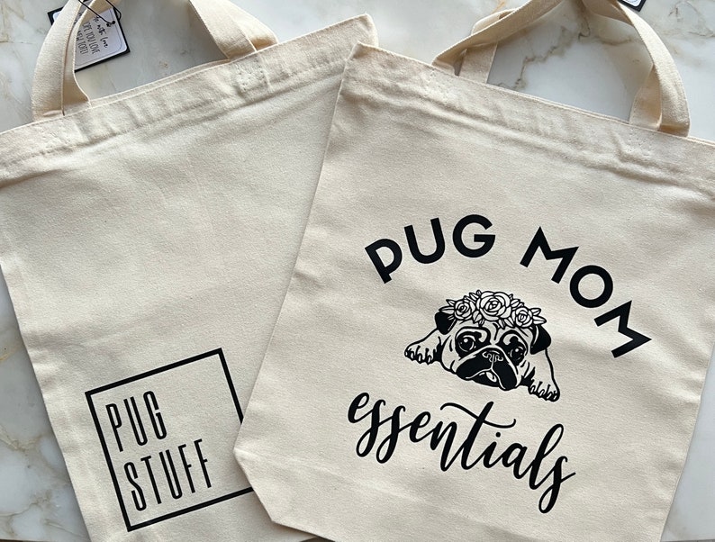 Op de afbeelding: Twee beige canvas tassen met zwarte tekst en afbeeldingen. E&eacute;n tas zegt "Pug Mom essentials" met een zwart-witte illustratie van een mops die een bloemenkrans draagt. De andere tas zegt "Pug Stuff" in een zwart vierkant.