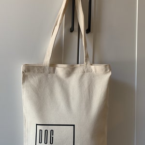 Op de afbeelding: Een beige canvas draagtas met zwarte handvatten en de tekst "DOG STUFF" gedrukt in een zwart vierkant aan de voorkant.