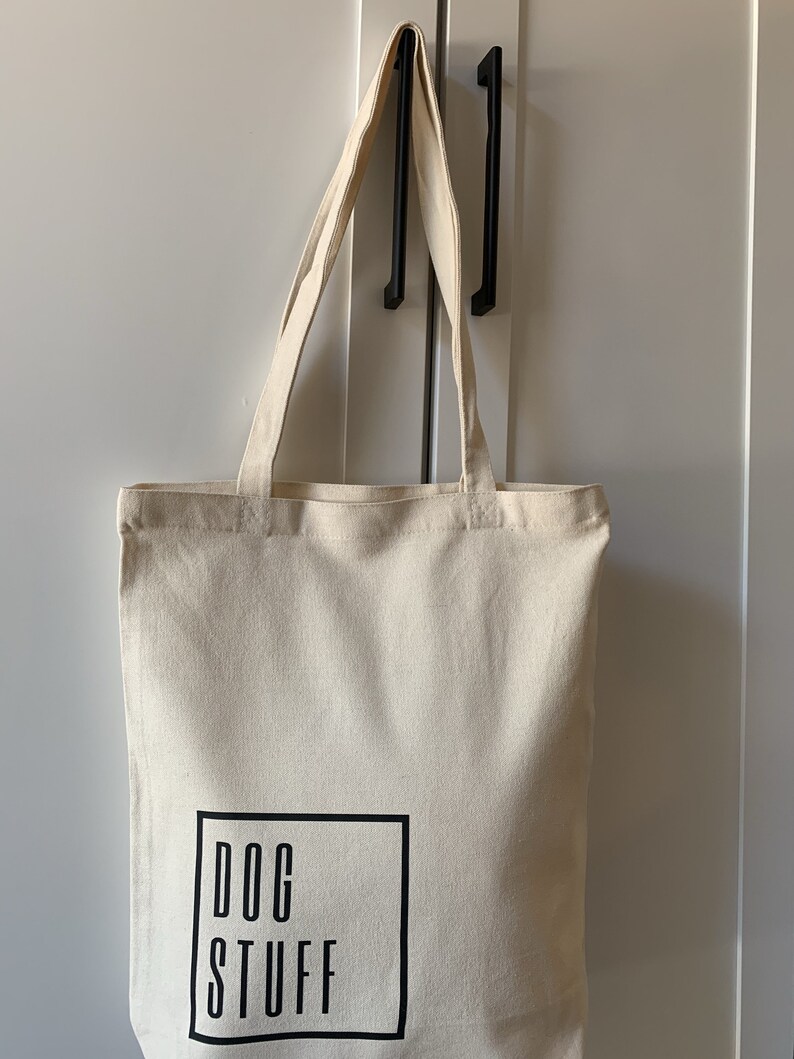 Op de afbeelding: Een beige canvas draagtas met zwarte handvatten en de tekst "DOG STUFF" gedrukt in een zwart vierkant aan de voorkant.
