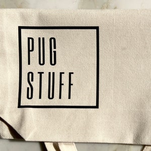 Op de afbeelding: Een witte canvas draagtas met zwarte banden en een zwart vierkant logo met de tekst "PUG STUFF" in hoofdletters.