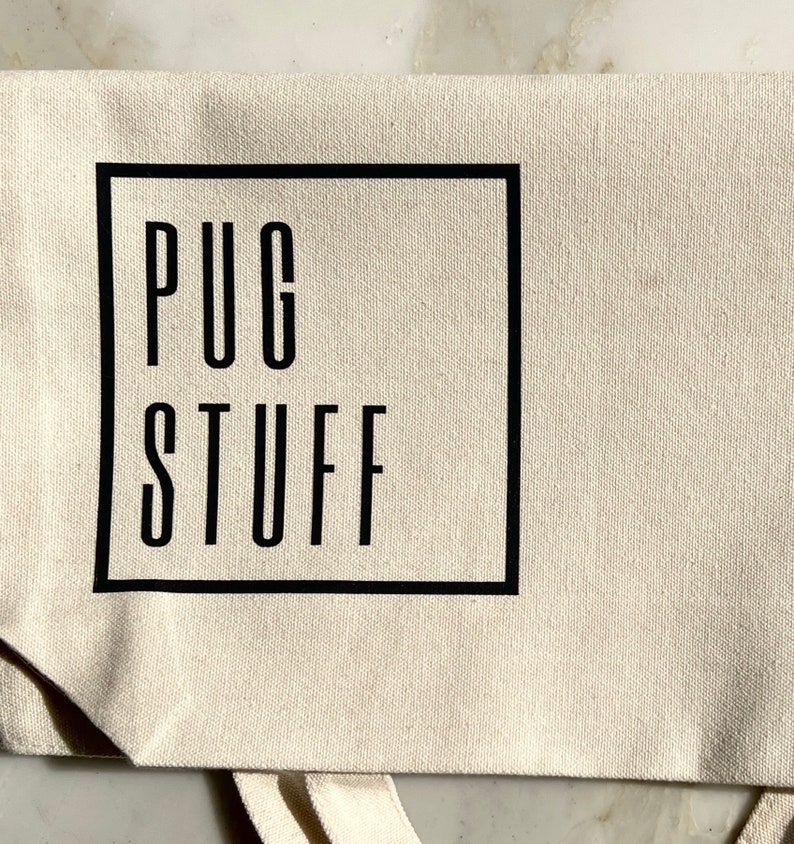 Op de afbeelding: Een witte canvas draagtas met zwarte banden en een zwart vierkant logo met de tekst "PUG STUFF" in hoofdletters.