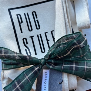 Op de afbeelding: Een witte canvas draagtas met zwarte tekst die "PUG STUFF" zegt en een groen-zwart geruit lint dat om de handvatten is gebonden. Een klein zwart-wit etiket met de tekst "JAX & OLLIE" is aan de tas bevestigd.
