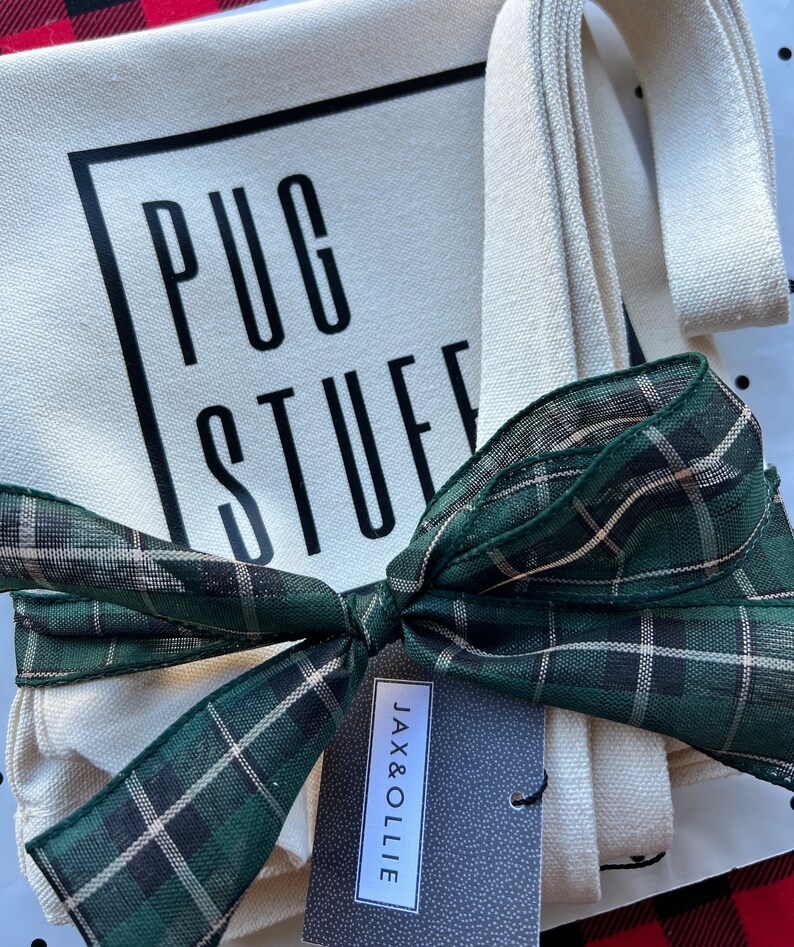 Op de afbeelding: Een witte canvas draagtas met zwarte tekst die "PUG STUFF" zegt en een groen-zwart geruit lint dat om de handvatten is gebonden. Een klein zwart-wit etiket met de tekst "JAX & OLLIE" is aan de tas bevestigd.