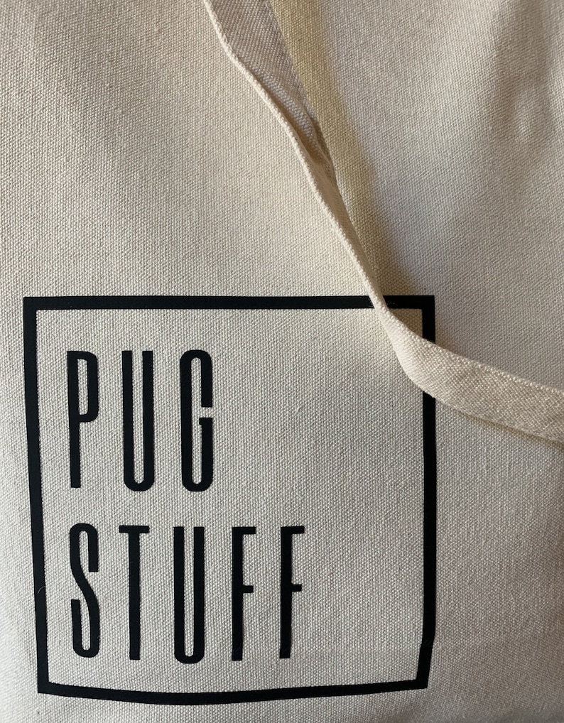Op de afbeelding: Een beige canvas draagtas met zwarte tekst die "PUG STUFF" leest in een zwart vierkant.