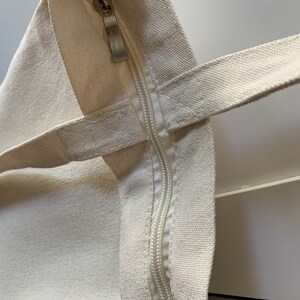 Op de afbeelding: Close-up van een cr&egrave;mekleurige canvas tote bag met een ritssluiting. De rits is gedeeltelijk open, waardoor de binnenkant van de tas zichtbaar is.