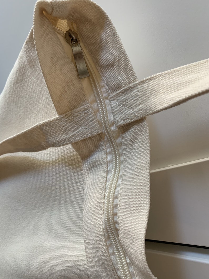 Op de afbeelding: Close-up van een cr&egrave;mekleurige canvas tote bag met een ritssluiting. De rits is gedeeltelijk open, waardoor de binnenkant van de tas zichtbaar is.