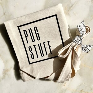 Op de afbeelding: Een witte canvas draagtas met zwarte banden en de tekst "PUG STUFF" gedrukt in een zwart vierkant.