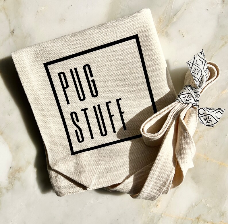 Op de afbeelding: Een witte canvas draagtas met zwarte banden en de tekst "PUG STUFF" gedrukt in een zwart vierkant.