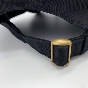 May include: Close-up of the back of a black baseball cap with a gold metal adjustable strap.
