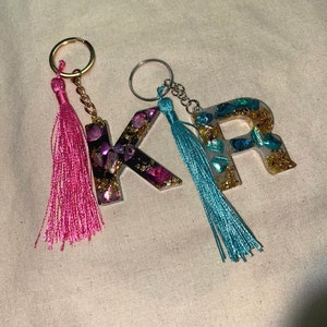 May include: Two resin letter charms with gold glitter and iridescent shell accents on gold keychains with pink and blue tassels.