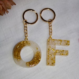 Peut inclure: Deux porte-clés dorés avec des breloques en résine blanche. Un charme est un cercle avec des paillettes dorées, l'autre est la lettre F avec des paillettes dorées.