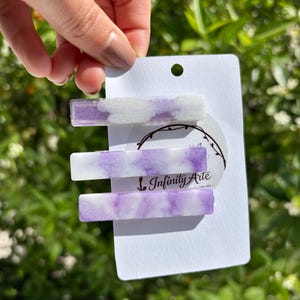 以下が含まれることがあります： 紫と白の大理石模様のデザインの長方形のヘアクリップが3つ。クリップは「Infinity Arte」の文字が入った白いカードに表示されています。背景は緑の葉のぼやけた景色です。