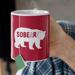 May include: A red ceramic mug with a white bear silhouette and the text "SOBEAR" printed on it. A green tea bag hangs from the rim of the mug.